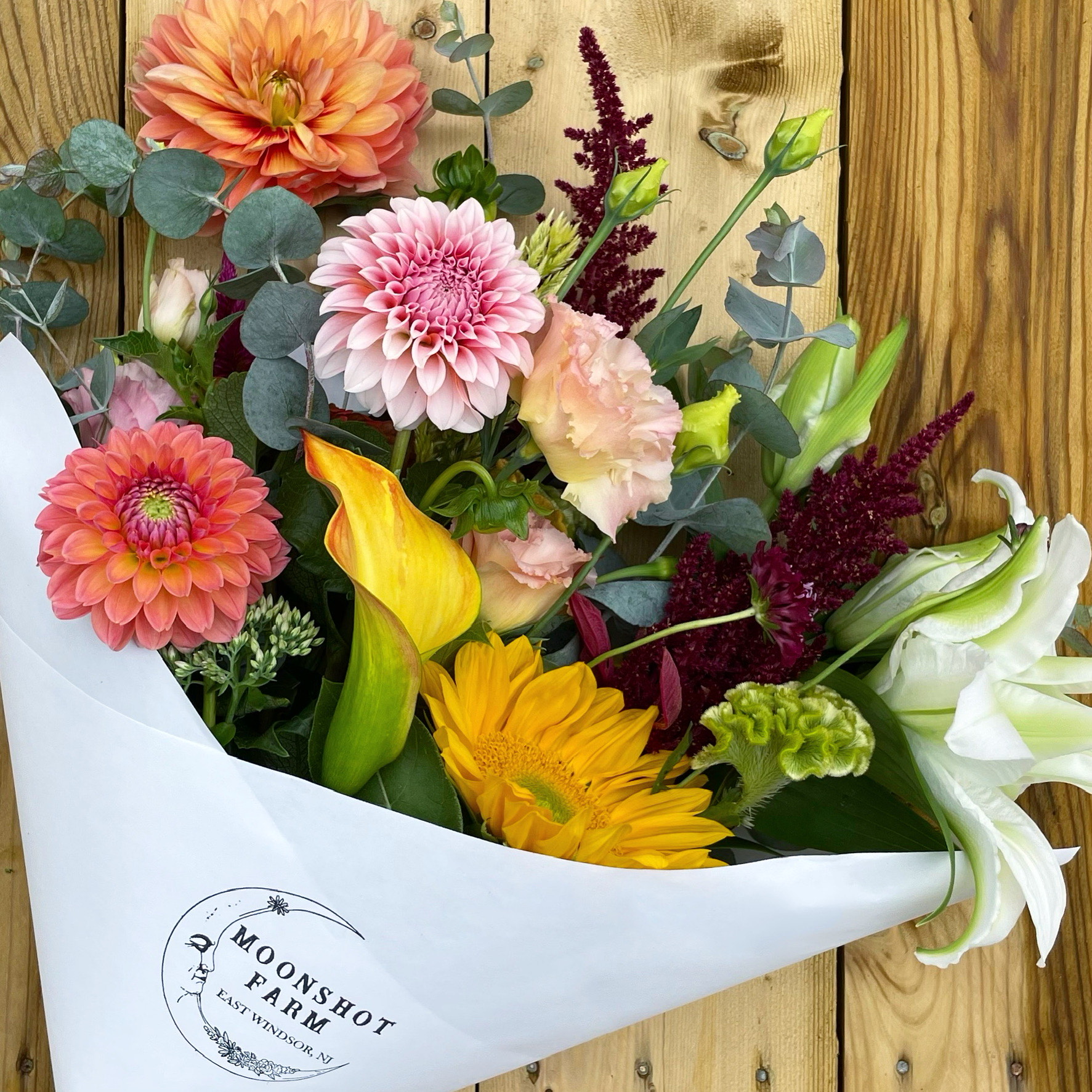 bouquet of pink, white, yellow and orange flowers wrapped in a white paper with a moon logo and the words Moonshot Farm